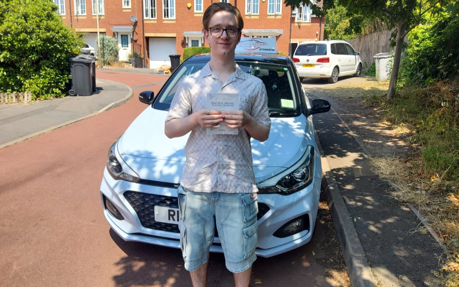 A learner holding his practical driving test pass certificate in front of the Rimit Sedani driving school car